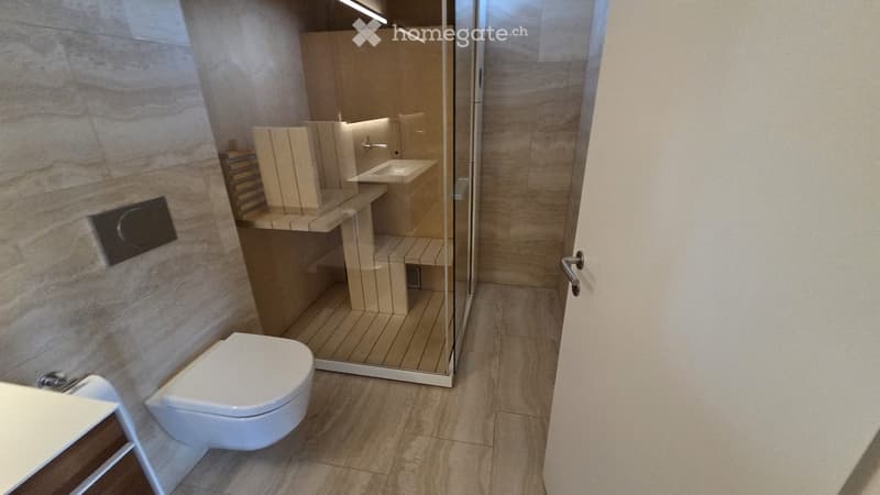 Modern bathroom with a toilet, sink, and glass shower enclosure. The walls are tiled in a light, textured pattern, and the floor is made of light-colored tiles.