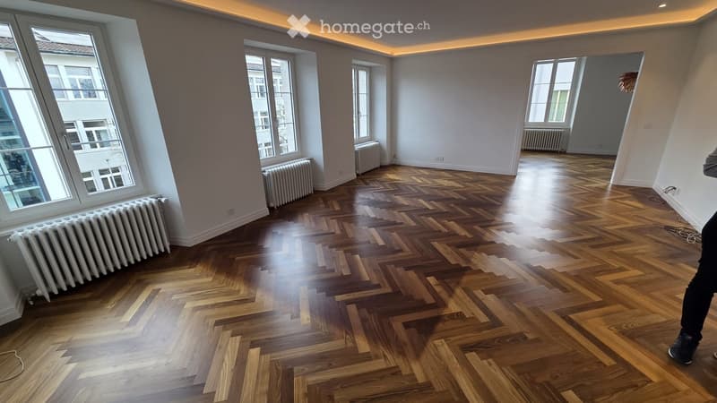 Empty room with wooden herringbone flooring, large windows, radiators, and white walls.