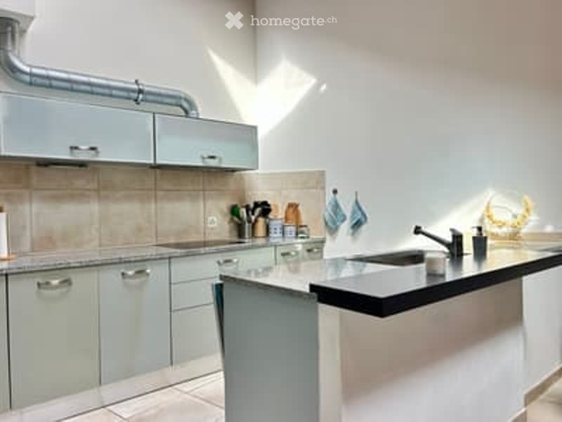 Modern kitchen with light grey cabinets, a black countertop, and a stainless steel sink. The space is bright with natural light and features a clean, minimalist design.