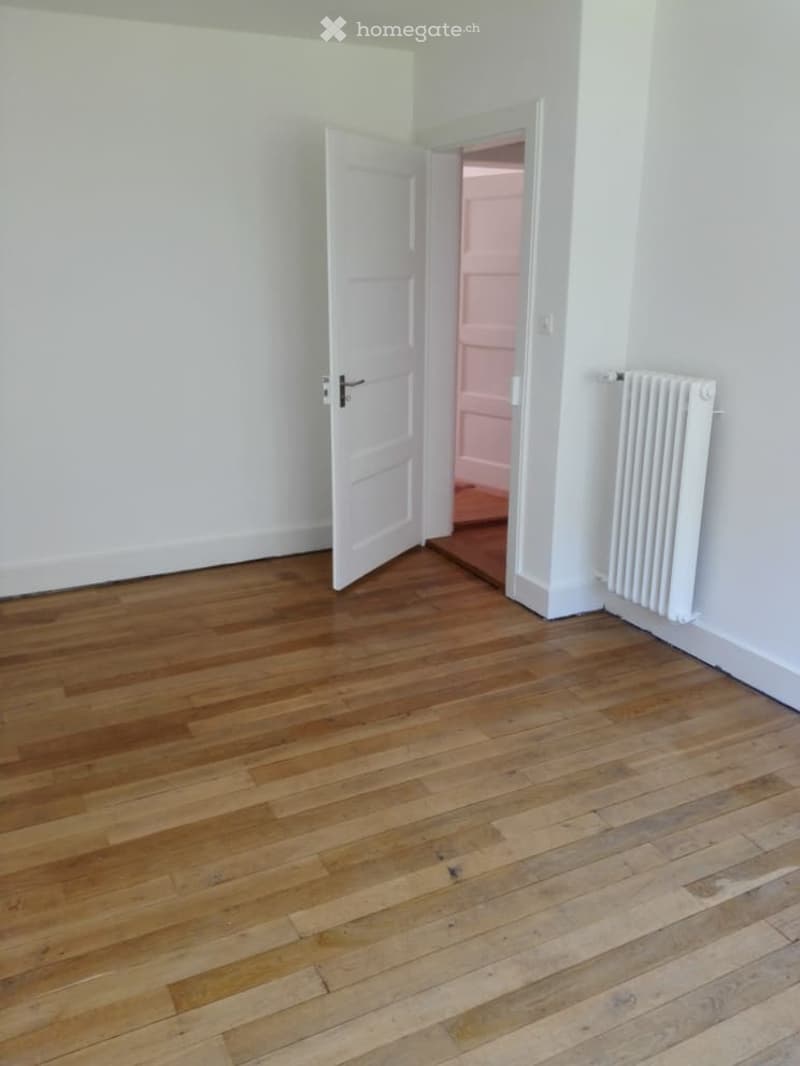 Empty room with wooden floor, open white door, white walls, white radiator