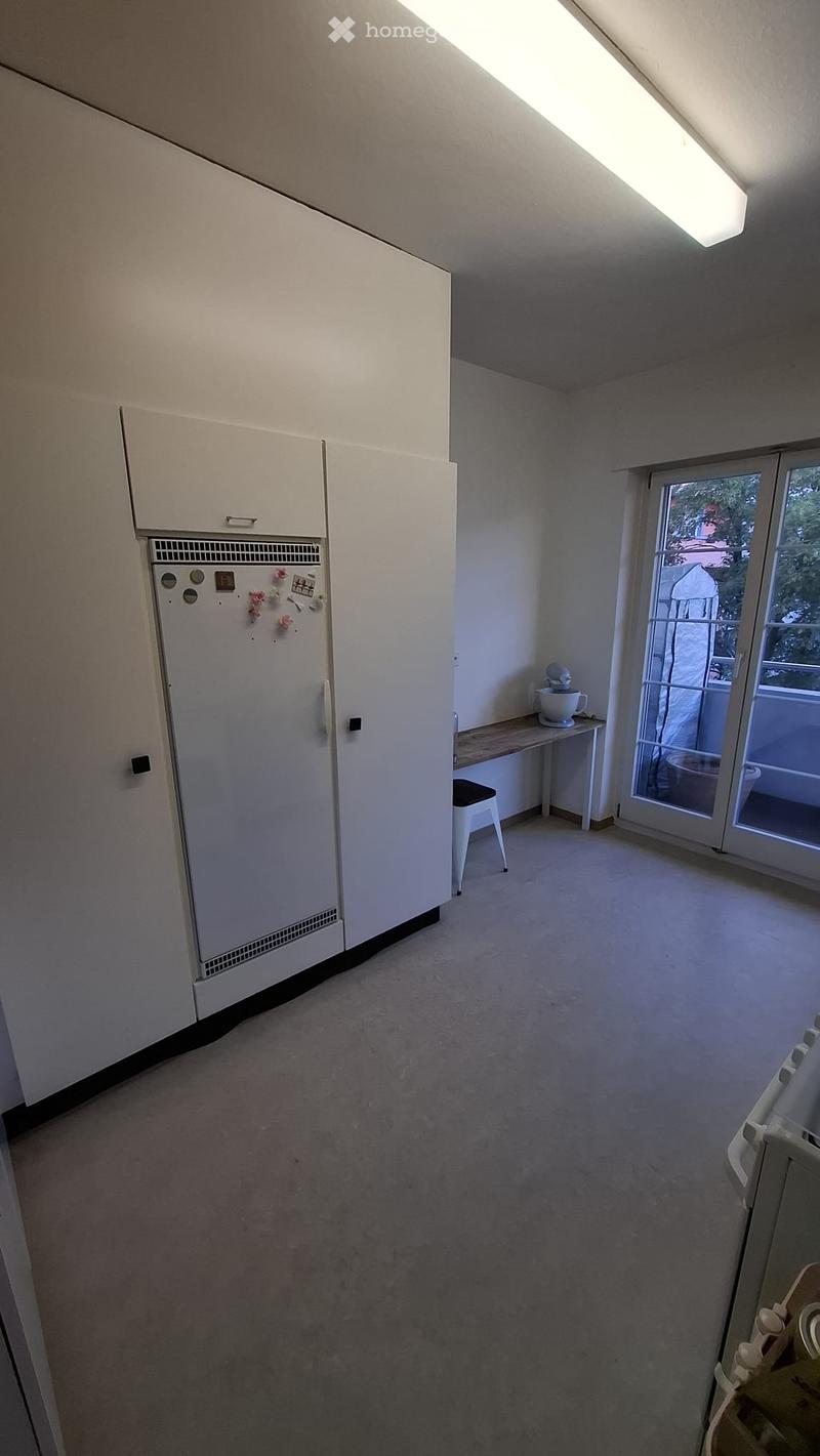 The room features a refrigerator, a small table with a mixer, and a stool near a sliding glass door leading to a balcony.