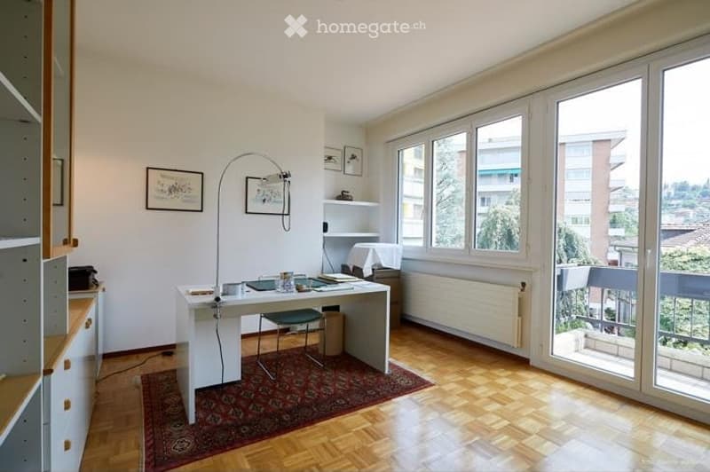 Bright, modern room with wooden flooring, large windows, and a balcony. Features a white desk, green chair, floor lamp, and picture frames on the wall.
