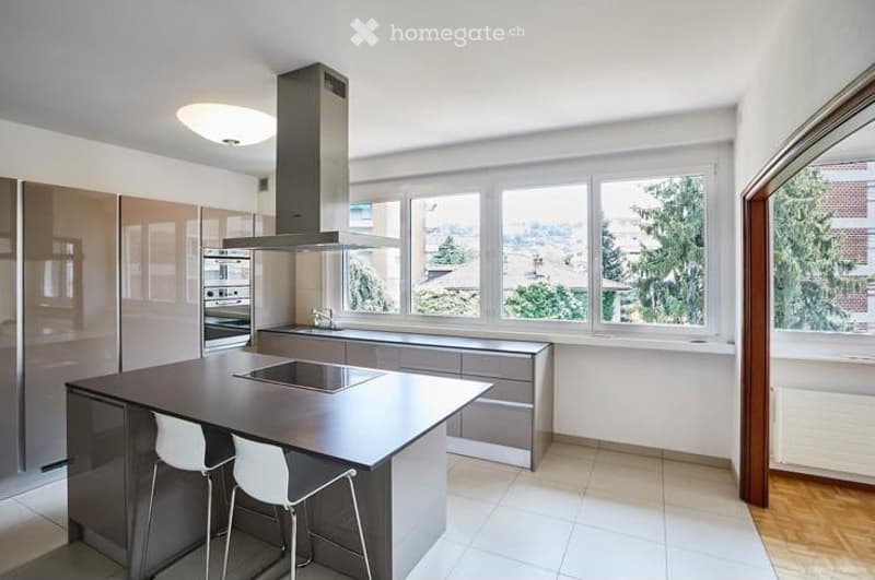 Modern kitchen with a central island, built-in stove, sink, and large windows offering a view of greenery outside.