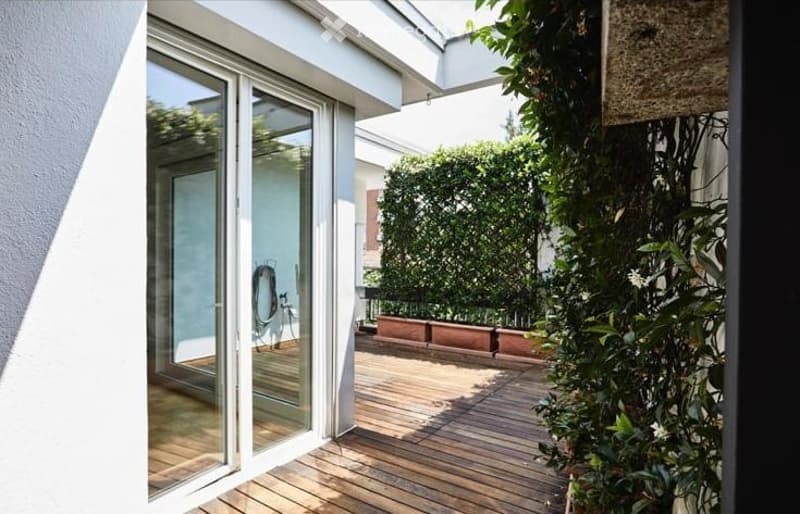 A modern wooden deck terrace with glass sliding doors leading to an indoor space, surrounded by greenery and potted plants.