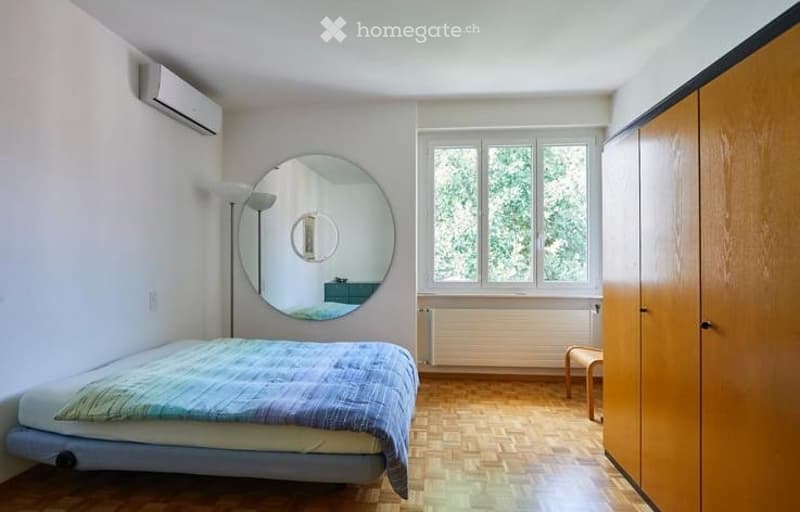 A modern, minimalist bedroom with a single bed, a large round mirror, a wooden wardrobe, a small stool, and a floor lamp. The room features light-colored walls, wooden flooring, and large windows with a view of greenery.