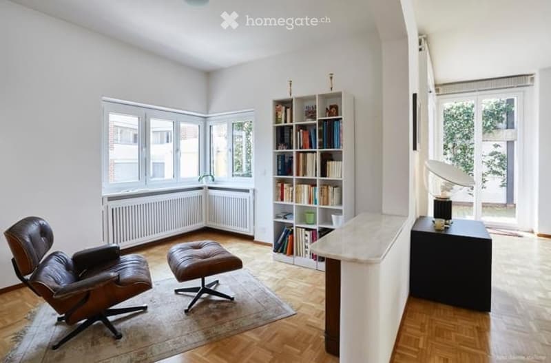 Bright, modern living room with large windows, wooden flooring, leather armchair, ottoman, bookshelf, and a small table with a lamp and cup.