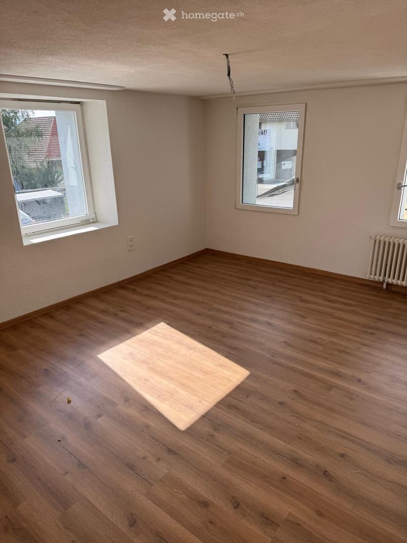 Empty room with wooden flooring, light-colored walls, and multiple windows, including one with a view of a neighboring building.
