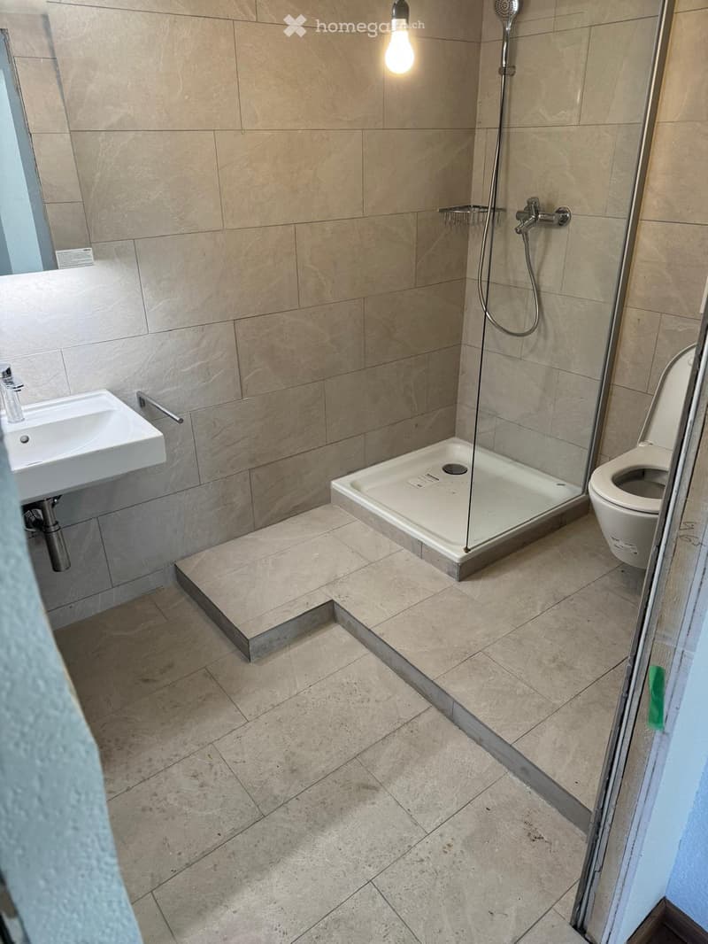 Modern bathroom with beige tiled walls and floor, featuring a white sink, toilet, and glass-enclosed shower stall.