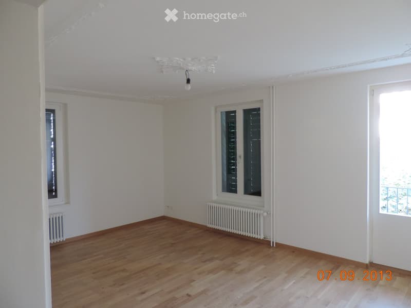 Spacious room with white walls, hardwood flooring, and a radiator. The room has large windows providing natural light.