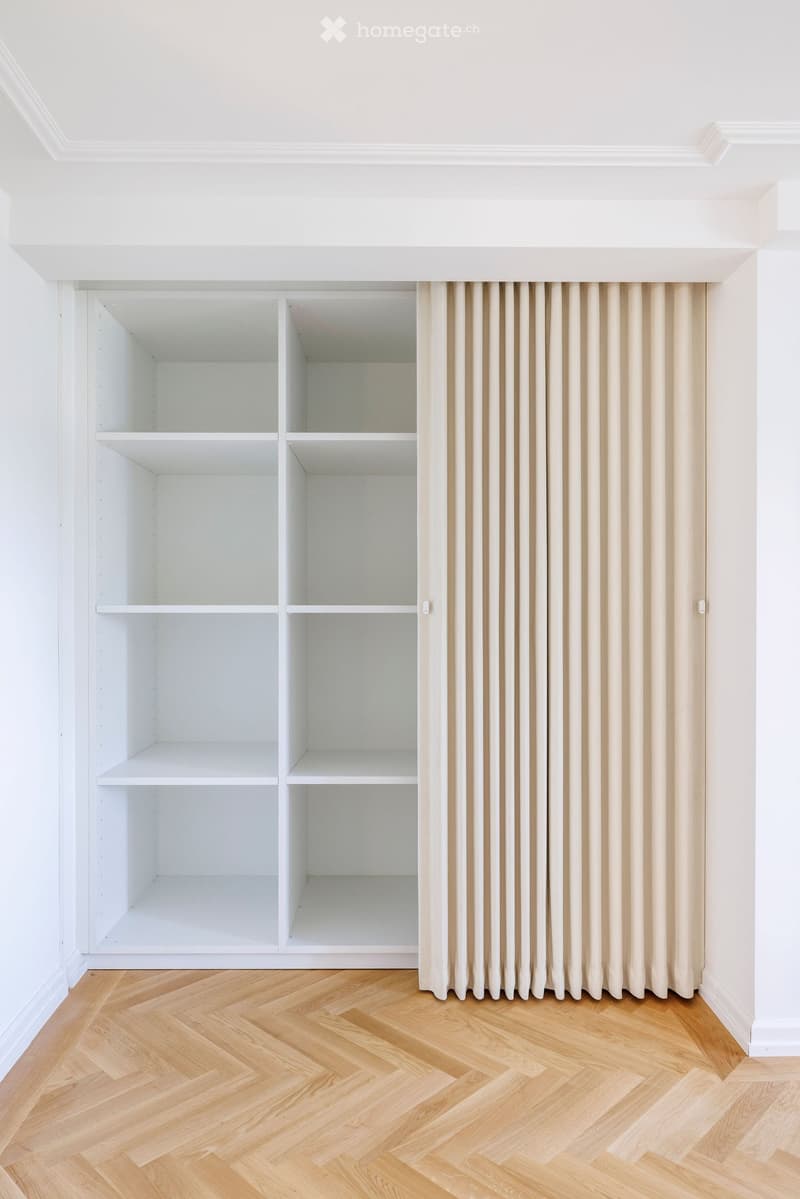 Empty room with white built-in shelving units and light wooden herringbone flooring. Beige curtains cover a window area.
