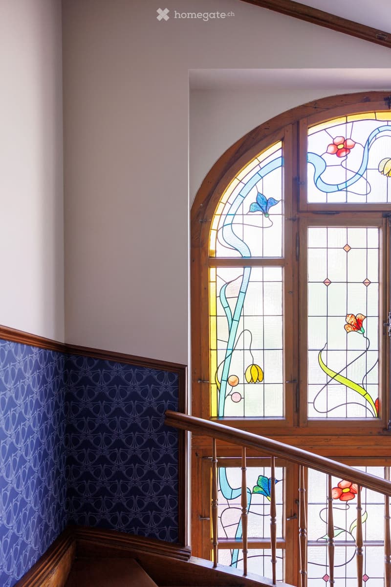 Staircase with wooden railings and stained glass windows featuring floral designs, blue patterned wallpaper on one wall.
