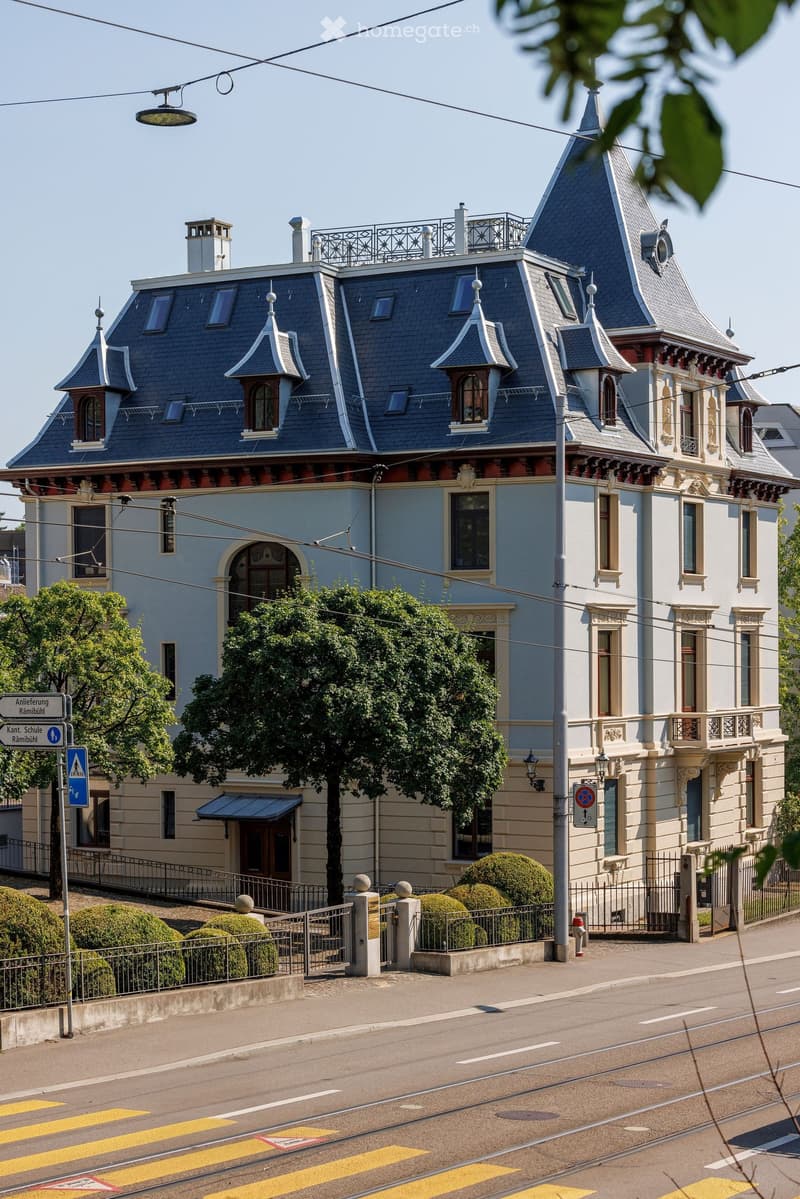 A large, ornate building with a steep, multi-pitched roof and decorative turrets. The facade is light blue with white trim and features arched windows and balconies. The building is surrounded by a low fence and well-maintained shrubs.