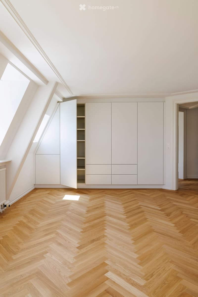Bright, empty attic room with wooden herringbone flooring, white walls, built-in white cabinetry, and skylight windows allowing natural light.