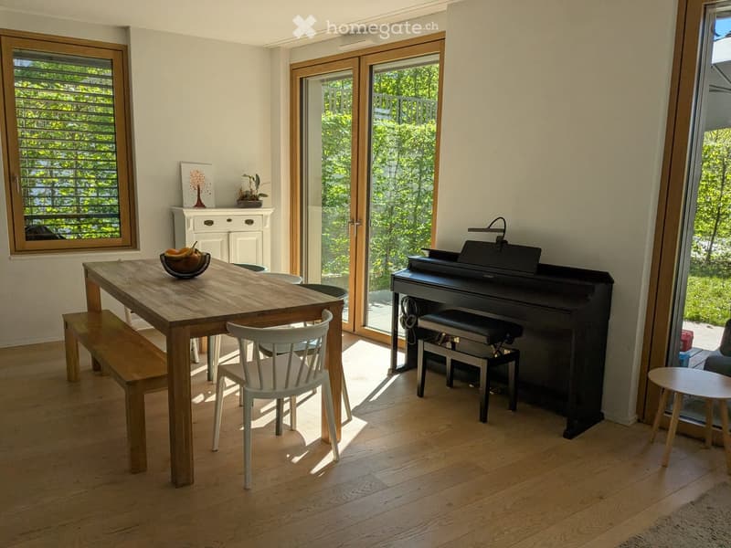 Dining room with wooden table, white chairs, wooden bench, piano, glass doors, wooden cabinet, potted plant, bowl of fruit, wooden floor, white walls
