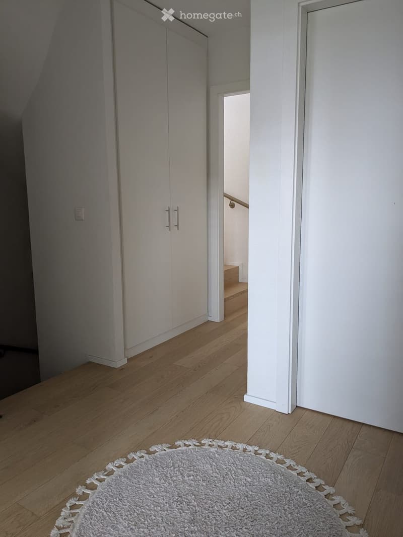 Empty room with wooden floor, white walls, white rug, large white wardrobe, and a staircase leading to another floor.