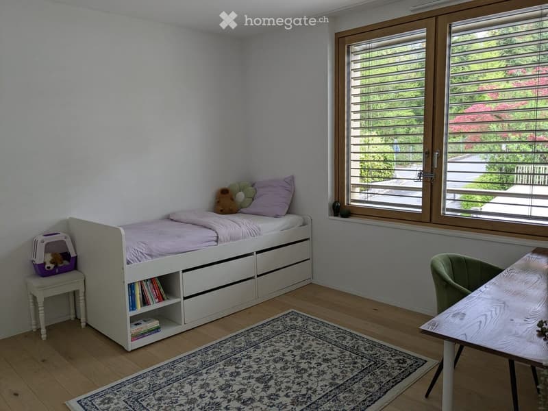 A small bedroom with a single bed, storage drawers, a small table with a pet carrier, a rug, and a window with blinds.