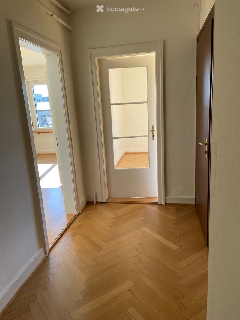 Empty hallway with herringbone-patterned wooden flooring, white walls, and two doors leading to adjacent rooms.