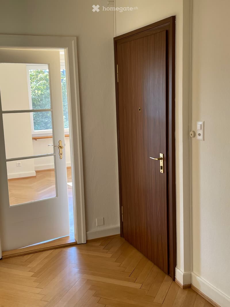 Hallway with wooden floor, white walls, and two doors: one solid wooden door and one glass-paned door leading to another room.
