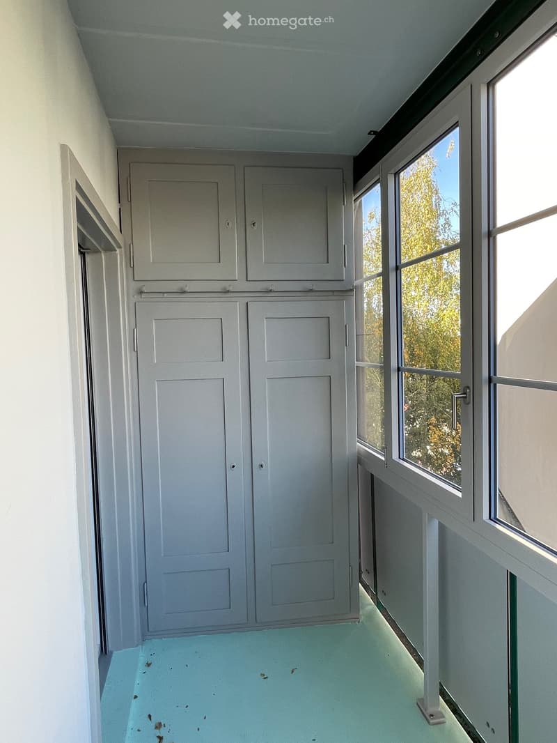 Empty room with light blue flooring, white walls, and large windows with a view of greenery outside.