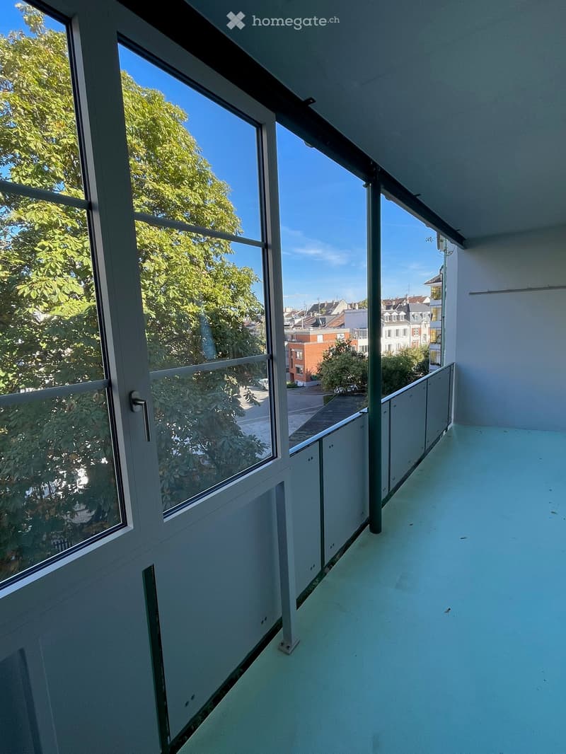Modern balcony with large windows, glass railing, and a view of trees and buildings.
