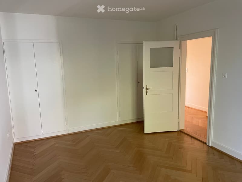 Empty room with wooden herringbone flooring, white walls, built-in white closet, and an open doorway leading to another room.