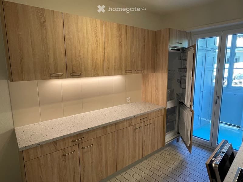 Modern kitchen with light wooden cabinets, granite countertop, and built-in appliances including a refrigerator and dishwasher. The space features tiled flooring and an open doorway leading outside.