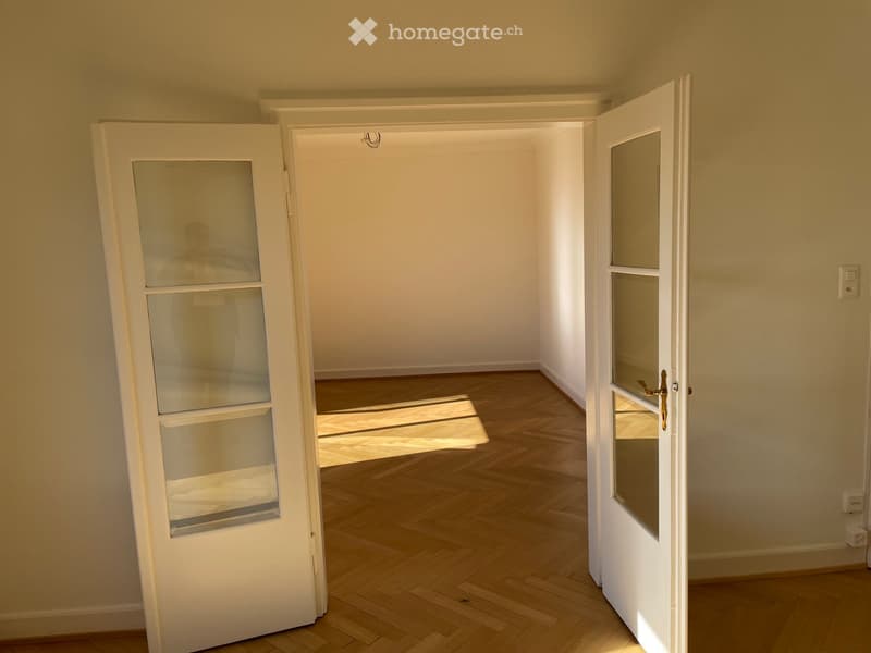 Empty room with wooden flooring and white walls, featuring two open doors leading to adjacent spaces.