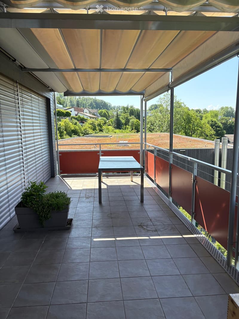 Balcony with tiled floor, covered by a retractable awning, featuring a small table and a potted plant, with a view of greenery and neighboring buildings.