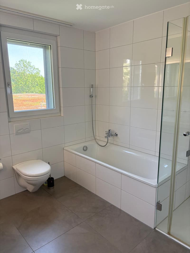 Modern bathroom with white tiles, bathtub, toilet, and glass-enclosed shower. Features a large window with a view of greenery outside.