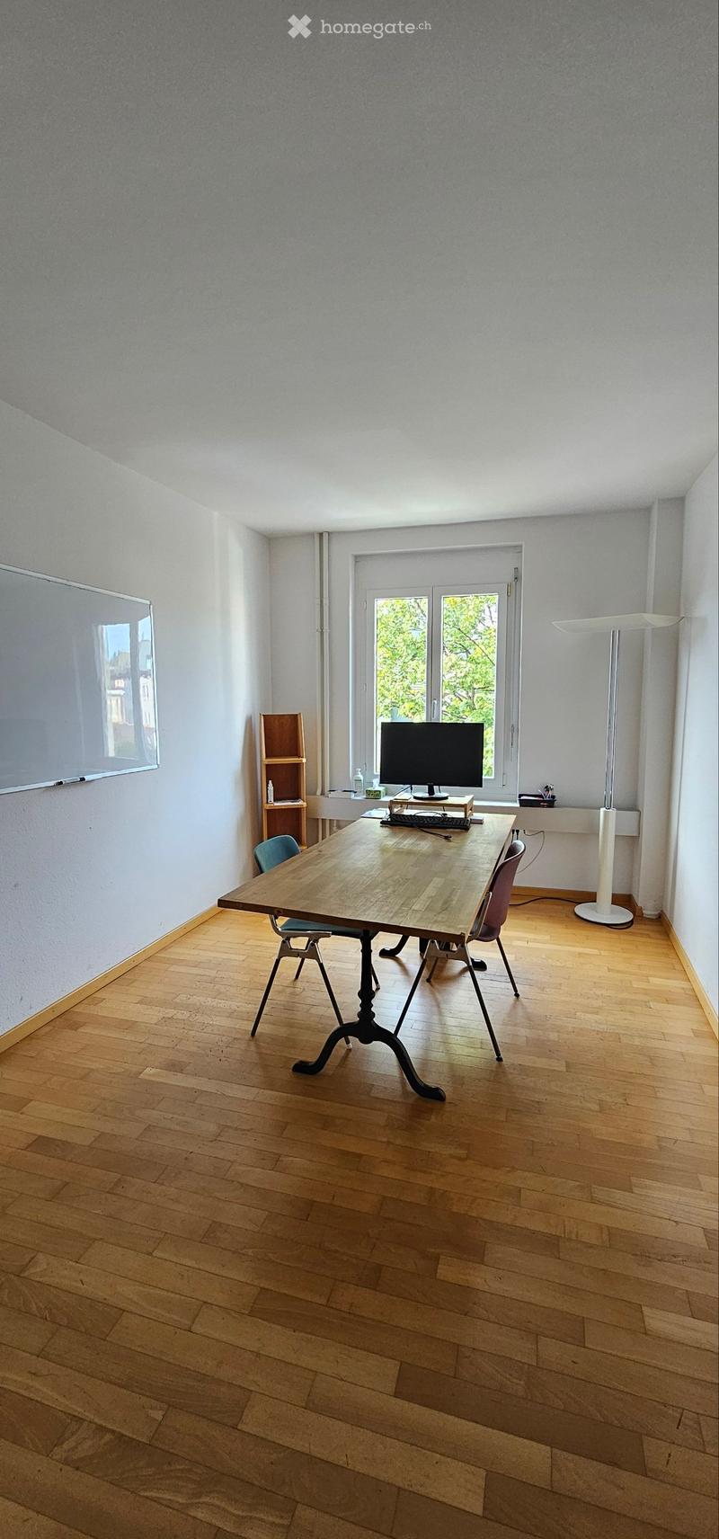 Bright room with wooden floor, large table, chairs, screen, keyboard, shelf, and floor lamp.