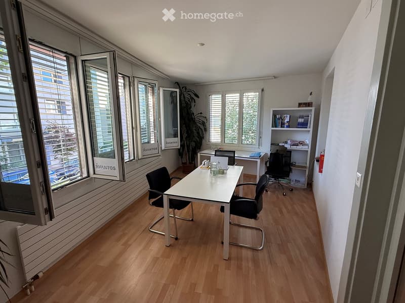 Modern office space with wooden flooring, large windows, a white desk, chairs, a laptop, and a bookshelf with books and decorative items.