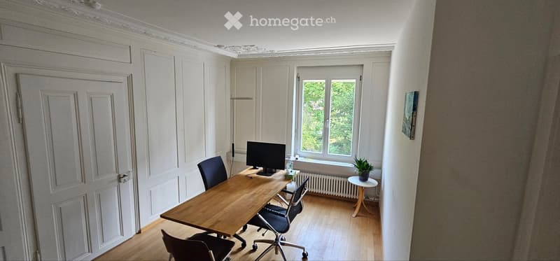 Bright room with wooden floor, large window, desk with computer, chairs, small side table with plant, and white paneled walls.