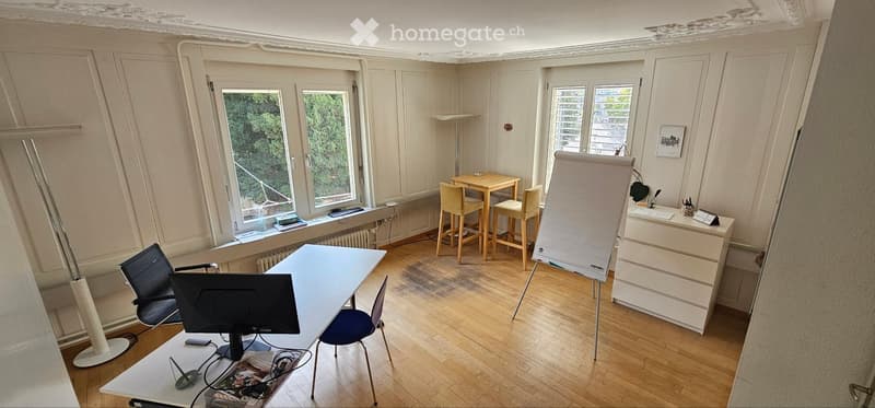 Office space with wooden flooring, large windows, a desk with a computer, chairs, a small table with stools, and a whiteboard.
