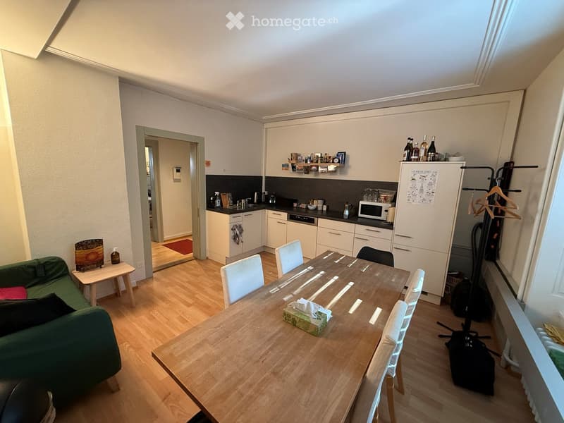 A modern, open-plan living and dining area with a green sofa, wooden dining table, white chairs, and a small kitchen area with cabinets, a refrigerator, and a microwave.