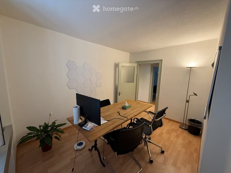 A modern, well-lit office space with a wooden desk, ergonomic chairs, a large screen, and a potted plant. The room features light-colored walls, wooden flooring, and a stylish geometric wall decoration.