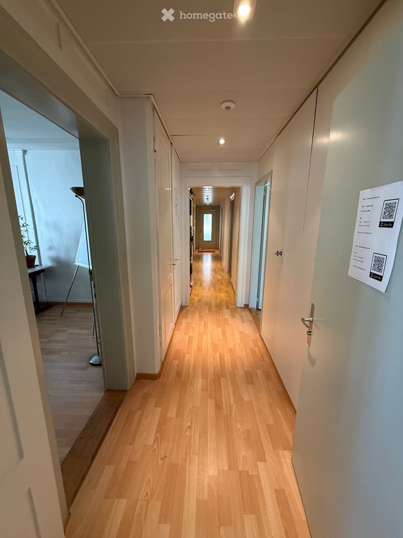 Long, narrow hallway with light wooden flooring, white walls, and several doors on both sides. A poster with a QR code is visible on the right wall.