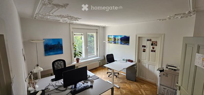Office space with wooden flooring, large window, two desks with computers, chairs, lamps, and artwork on the walls.