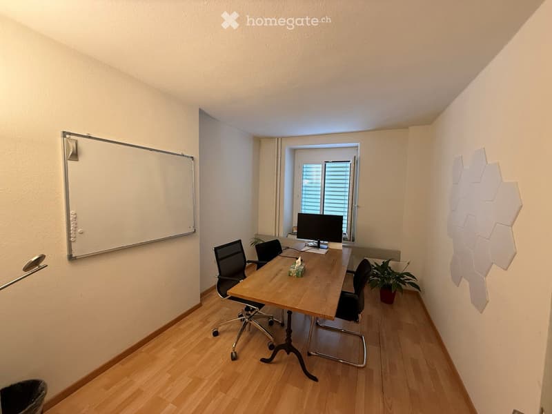 Office space with wooden floor, whiteboard, desk, chairs, computer screen, and small plant.