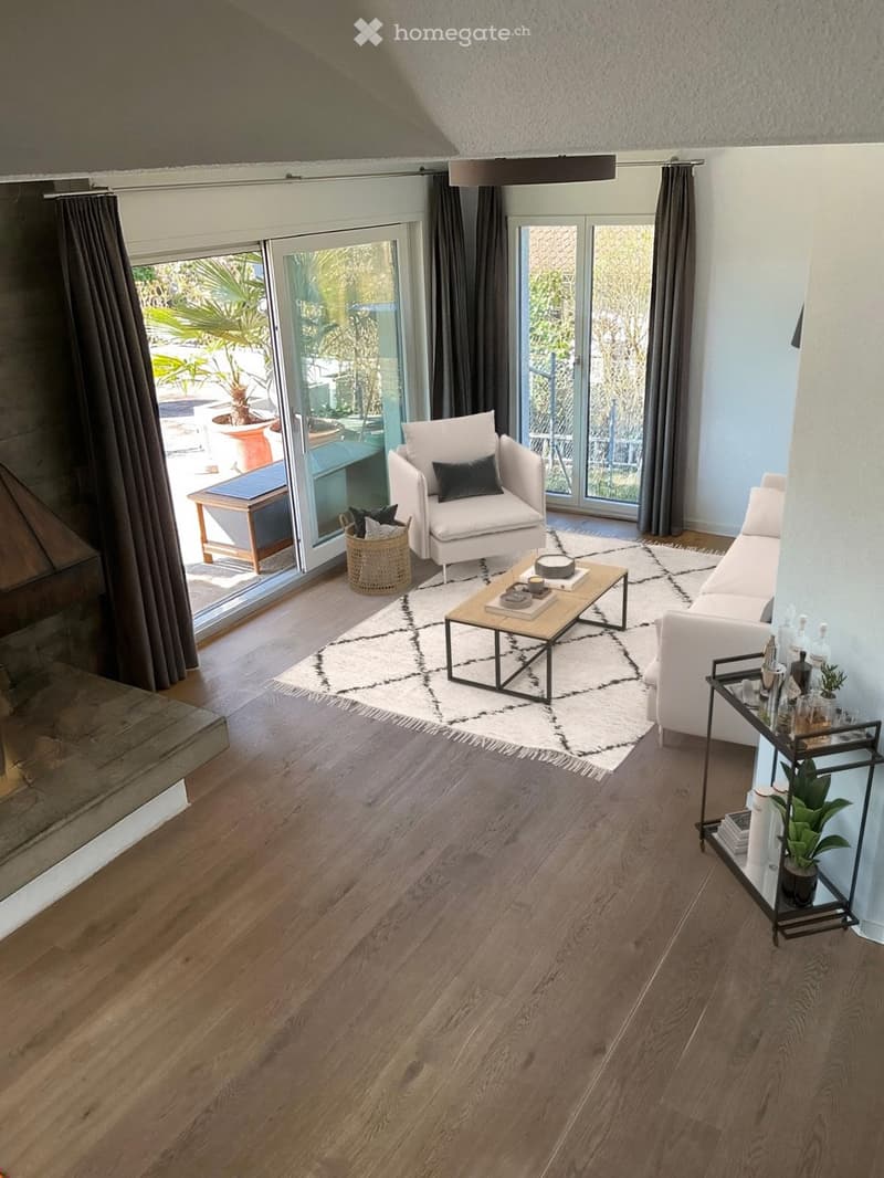 Living Room. Modern living room with wooden flooring, large windows, and a view of a garden. Features a white sofa, armchair, coffee table, side table, and a sideboard with bottles and a plant.