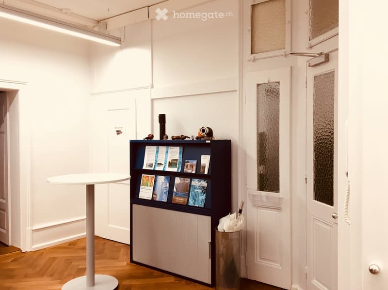 Small, modern, white room with wooden floor, a white round table, a small bookshelf, and a trash can.