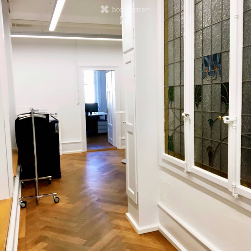 A hallway with white walls, wooden floors, and a view of an open room with a desk and monitor