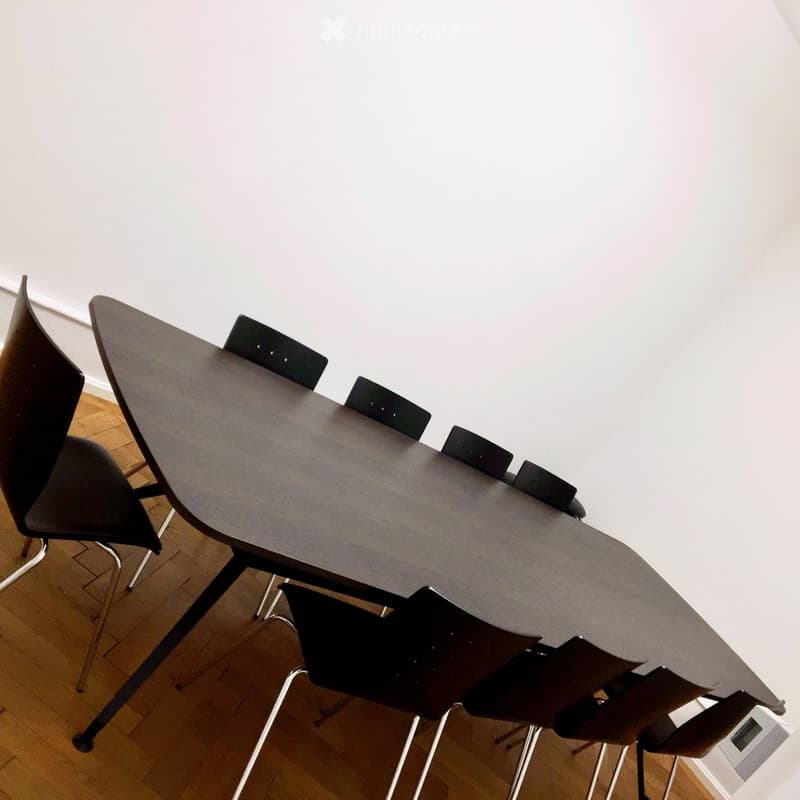 a large, oval-shaped, dark-colored wooden table in a room with a white wall and a wooden floor, with six black chairs around it.