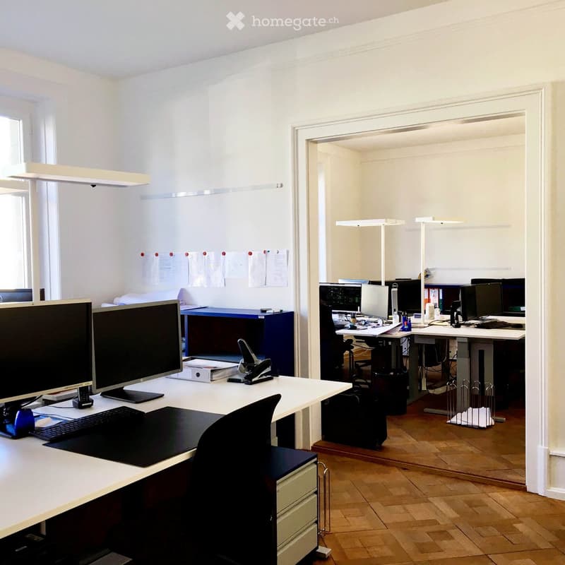 white walls, wooden floor, office desks with computer, chairs, monitors, keyboard, mouse, drawers, overhead lights
