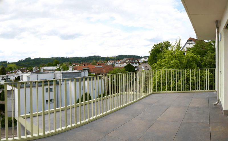 Balcony with tiled floor and metal railing, overlooking a town with houses, trees, and distant hills.