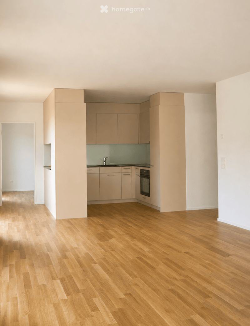 Empty room with light wooden flooring, white walls, and a small kitchen area with cabinets and appliances.