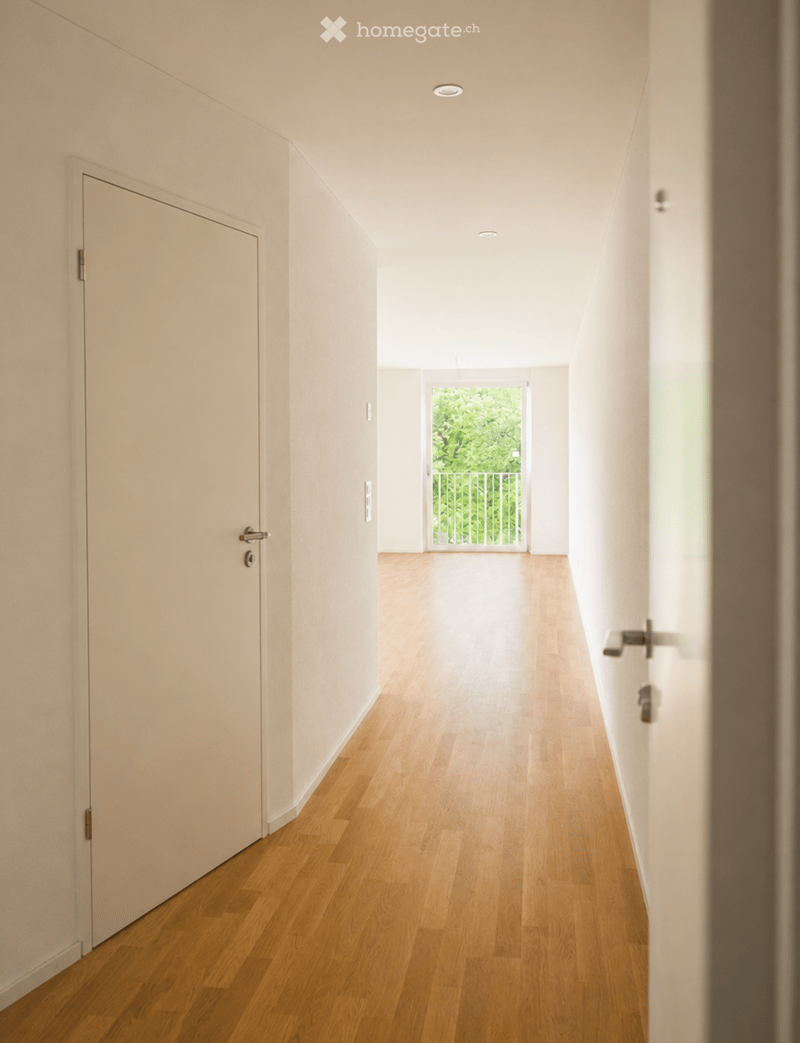 A modern, minimalist hallway with light wooden flooring, white walls, and recessed ceiling lighting. Features a closed door on the left and an open doorway leading to a balcony or outdoor area with greenery visible beyond.