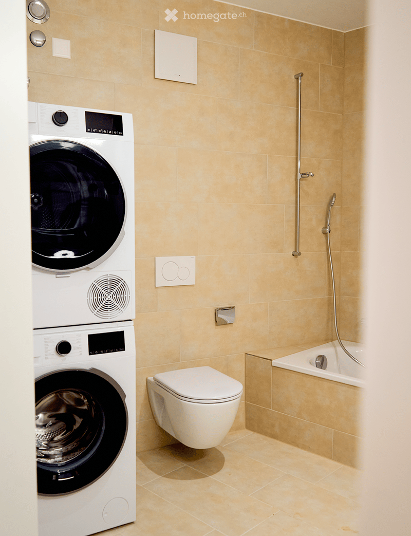 Modern bathroom with beige tiles, a white toilet, a bathtub with a shower hose, and a stacked washing machine/dryer unit.