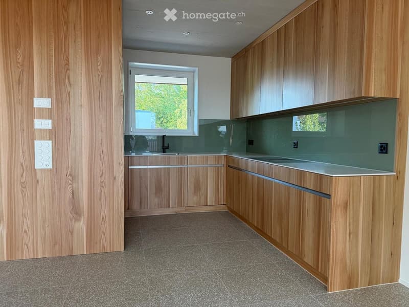 Modern kitchen with wooden cabinets, a central island, and a large window offering a view of greenery.