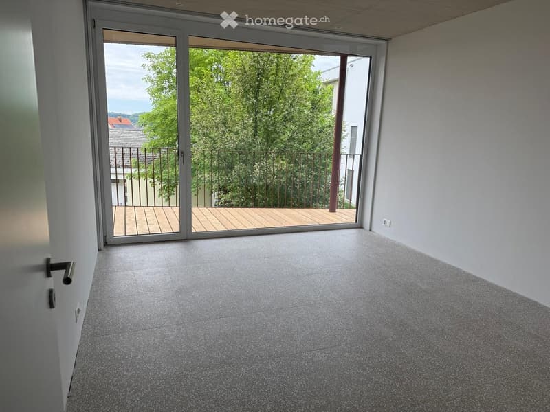 Empty room with grey tiled floor, white walls, and large sliding glass doors leading to a wooden balcony with metal railing and greenery visible outside.