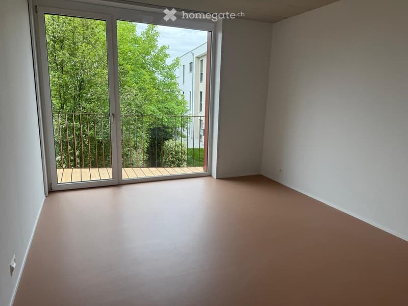 Empty room with light-colored walls and floor, large sliding glass doors leading to a balcony with wooden decking and metal railing.